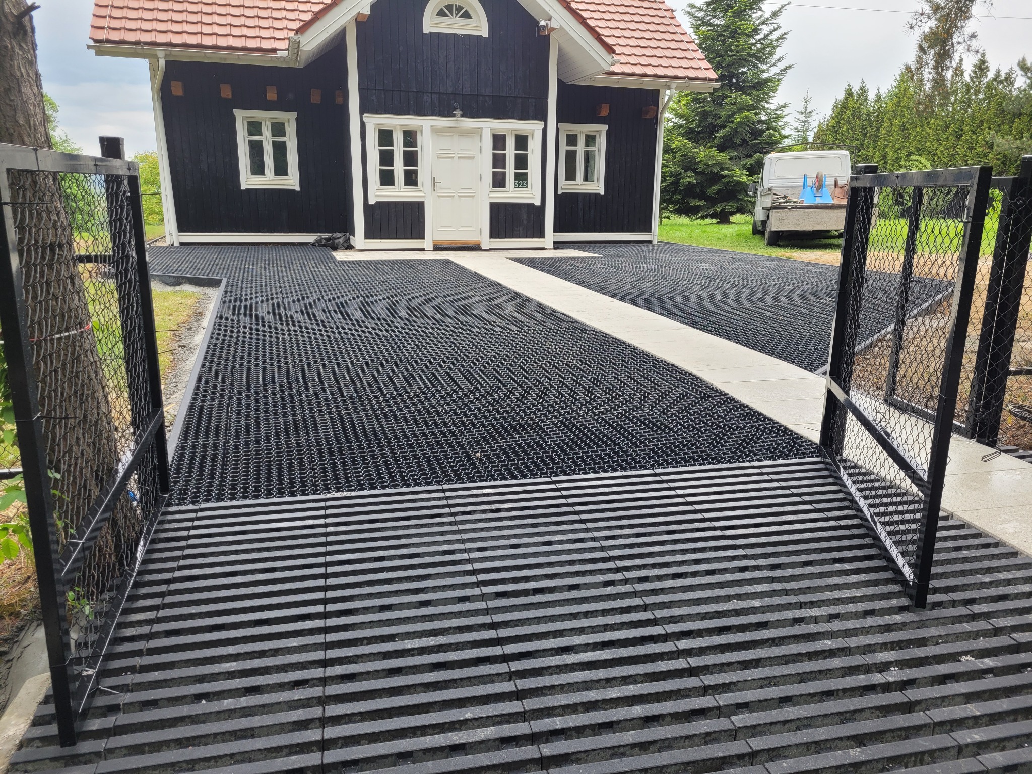 Nowoczesny podjazd z ażurową kostką i otwartą bramą, prowadzący do czarnego domu z białymi oknami i czerwonym dachem. W tle zaparkowany biały samochód dostawczy.