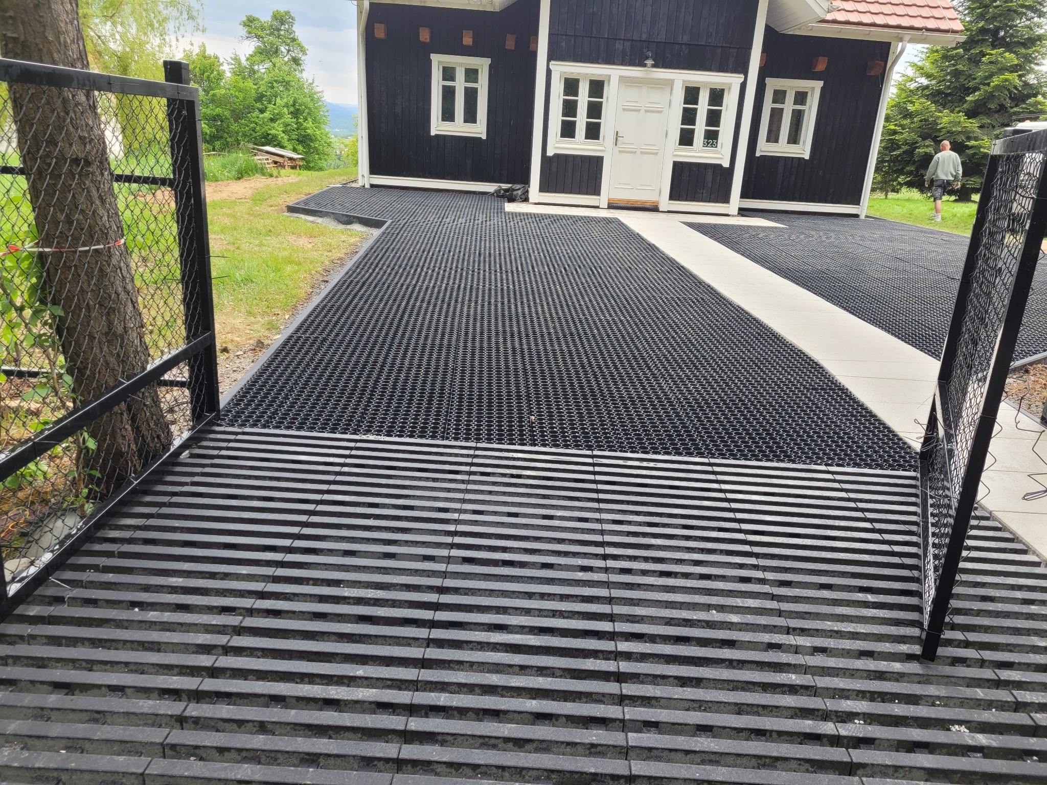 Ułożona, czarna nawierzchnia z kratki na podjeździe do domu z białymi oknami i drzwiami, widoczna brama z siatki i drzewo. W tle zieleń i osoba.