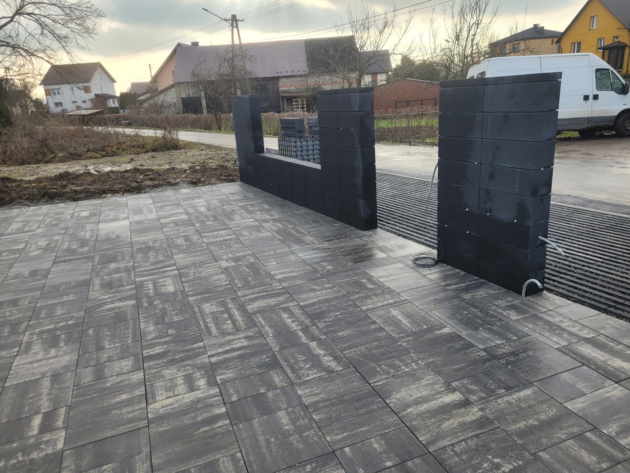 Ułożona szara kostka brukowa z elementami ogrodzenia z betonowych bloczków. Widoczny fragment drogi i zabudowania w tle. Świeżo po deszczu, mokra nawierzchnia.