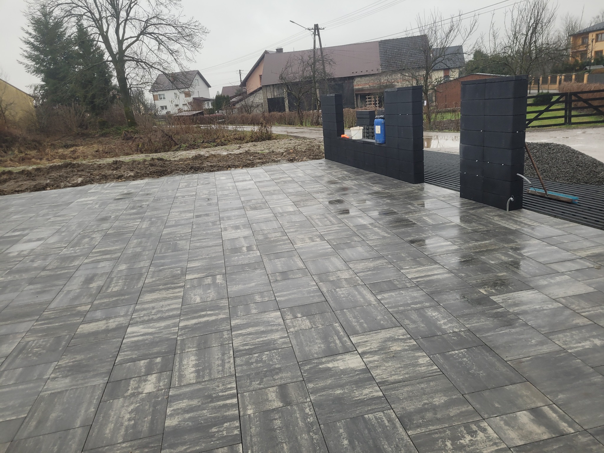 Ułożona, szara kostka brukowa w Tęgoborze, tworząca wzór. W tle widoczne elementy ogrodzenia z betonowych bloczków i przygotowany teren pod dalsze prace.