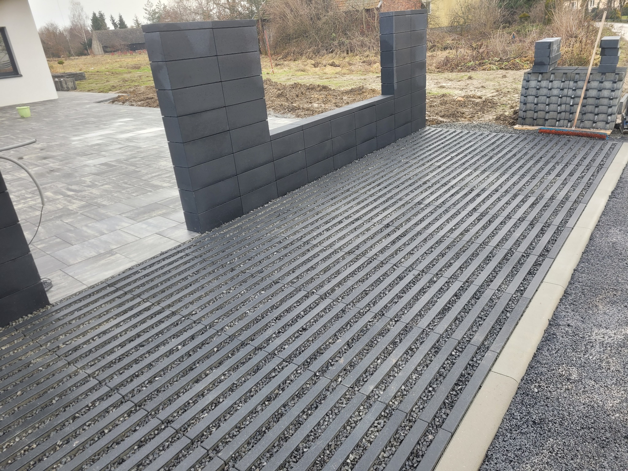 Szare ażurowe płyty z wypełnieniem żwirowym tworzą wzór na podjeździe. W tle fragment ogrodzenia z szarej kostki i ułożone płyty. Widoczny fragment domu i trawnik.