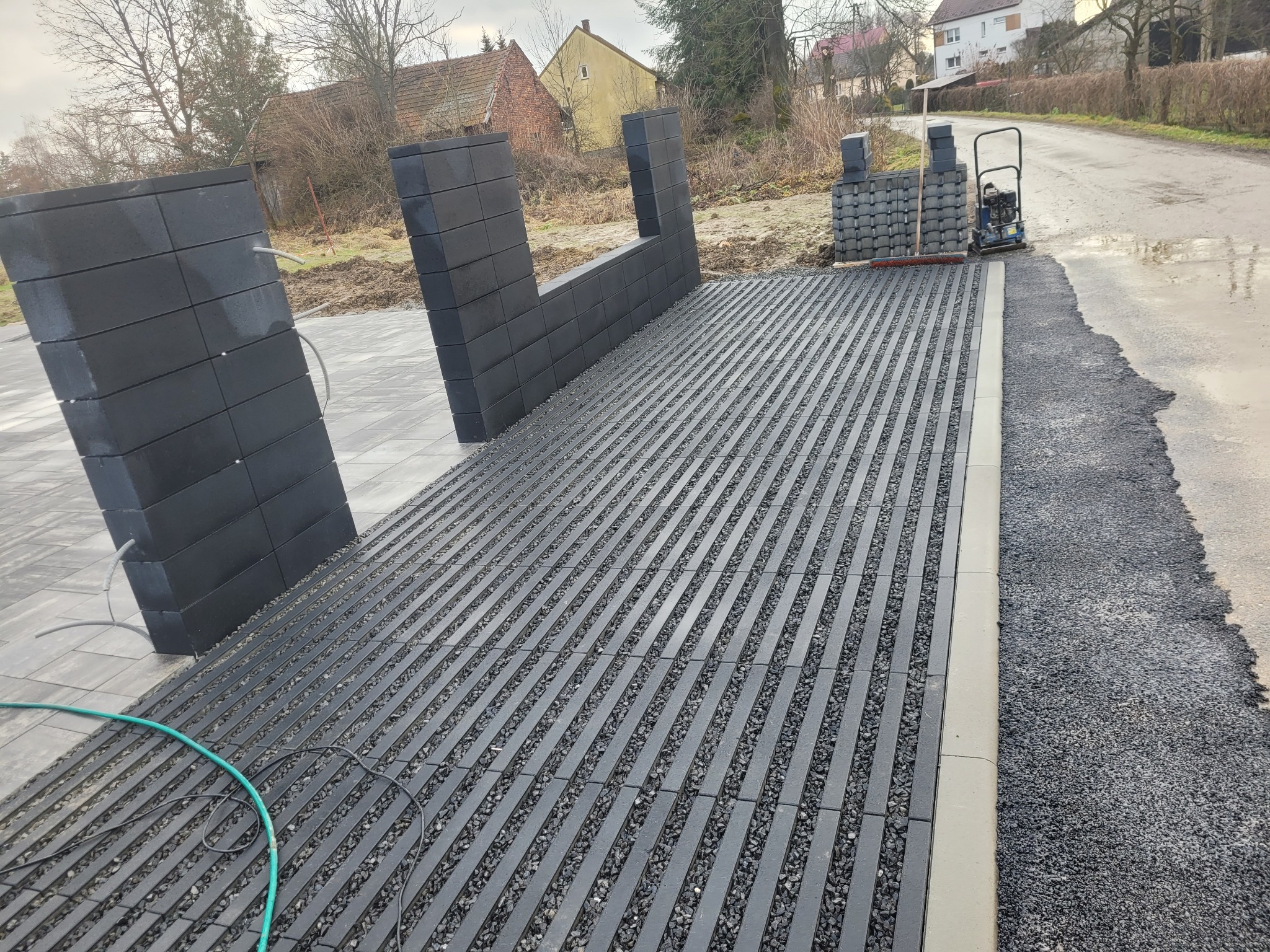 Nowoczesna nawierzchnia z szarych elementów betonowych i kamieni, częściowo ułożona, z widocznymi słupkami ogrodzeniowymi i narzędziami brukarskimi w tle.