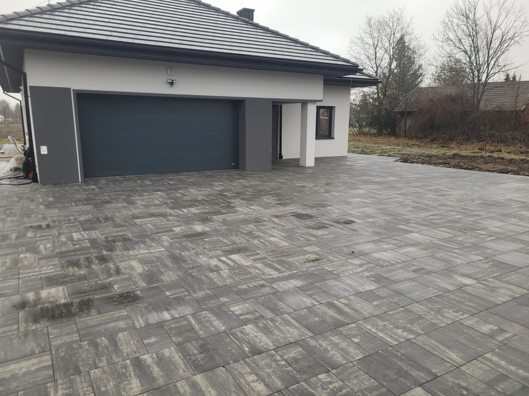 Nowoczesny dom z garażem i podjazdem wyłożonym szarą kostką brukową o nieregularnym wzorze. Widoczne detale wykończenia domu i otoczenie w jesiennej scenerii.