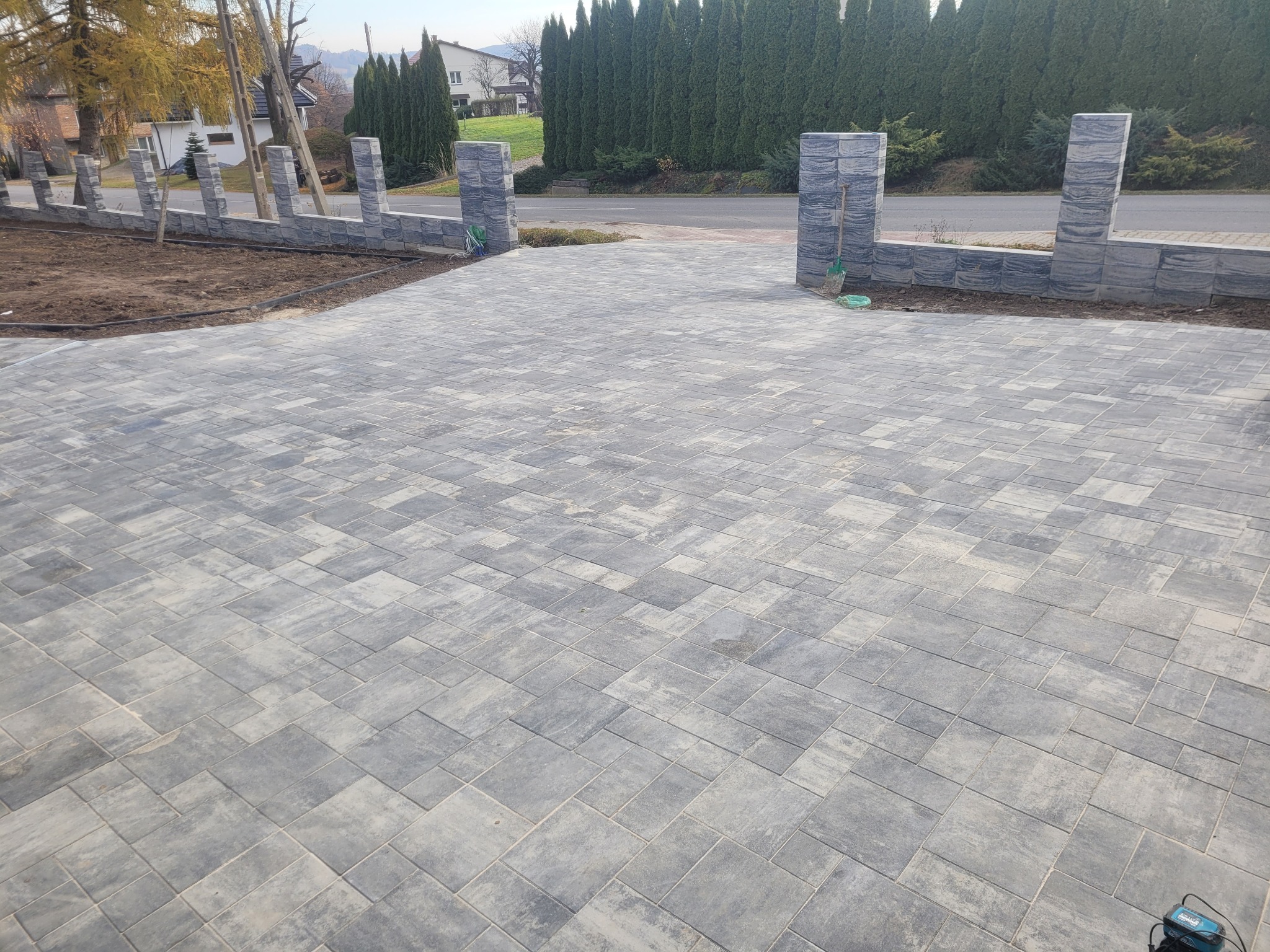 Ułożona szara kostka brukowa na podjeździe z elementami ogrodzenia z tego samego materiału. Widoczne narzędzia brukarskie. Tło: zieleń i zabudowa.