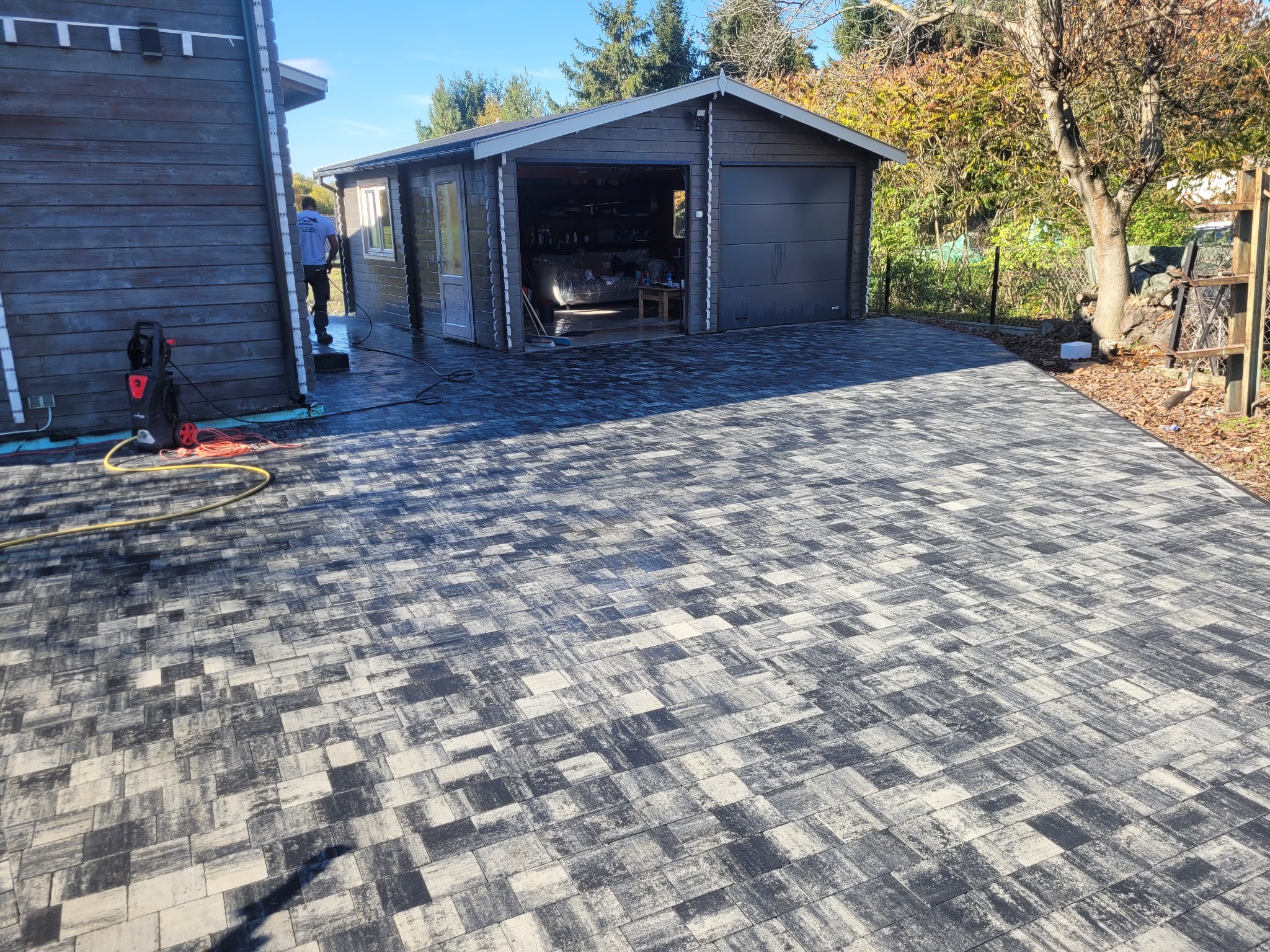 Nowa, szaro-biała kostka brukowa prowadząca do garażu z drewnianą elewacją. W tle widoczny pracownik z myjką ciśnieniową. Jesienne otoczenie.