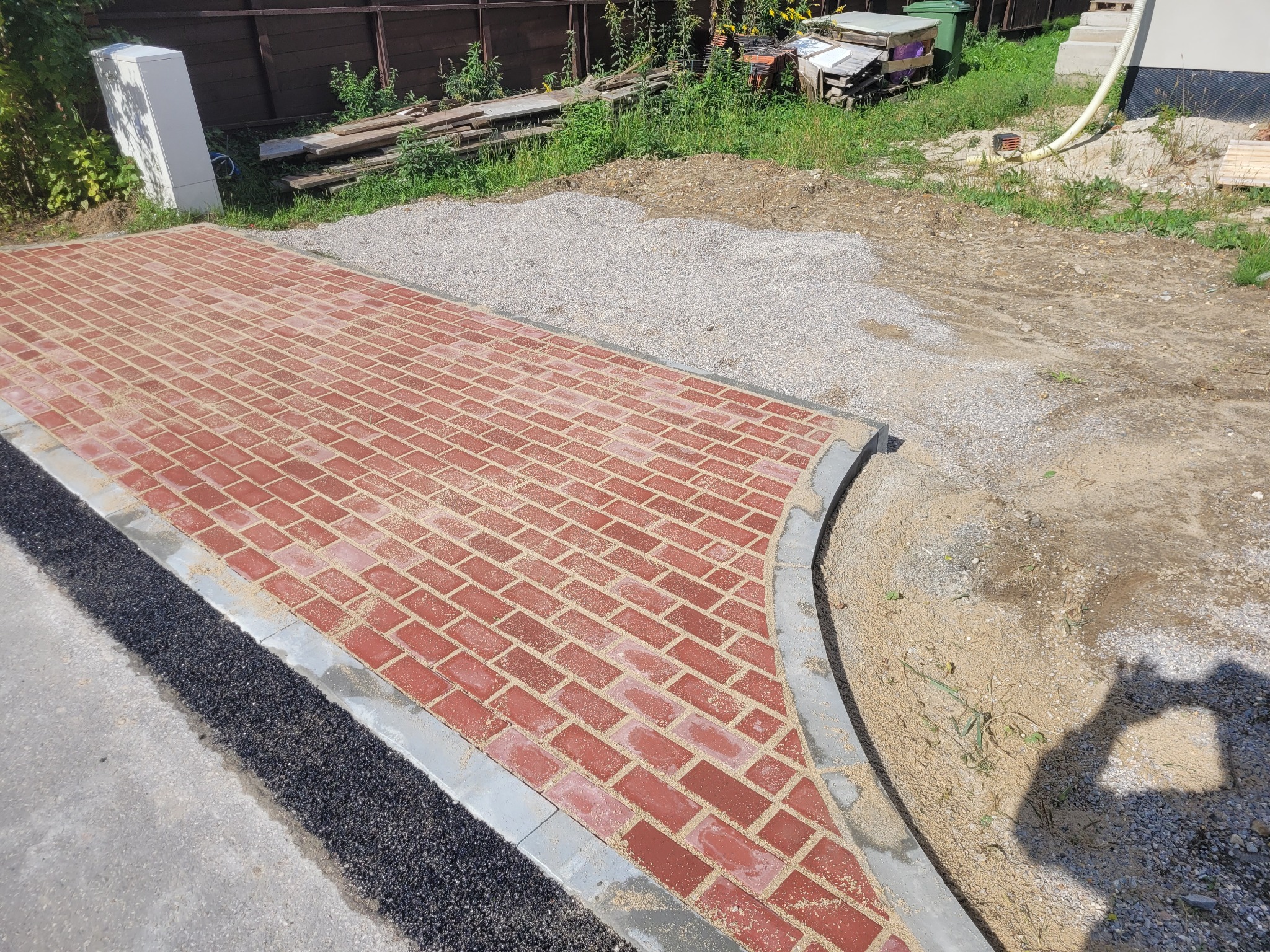 Czerwona kostka brukowa ułożona w jodełkę, obrzeża z szarego betonu, widoczne przygotowane podłoże z kruszywa i asfaltu. Świeżo wykonana ścieżka.
