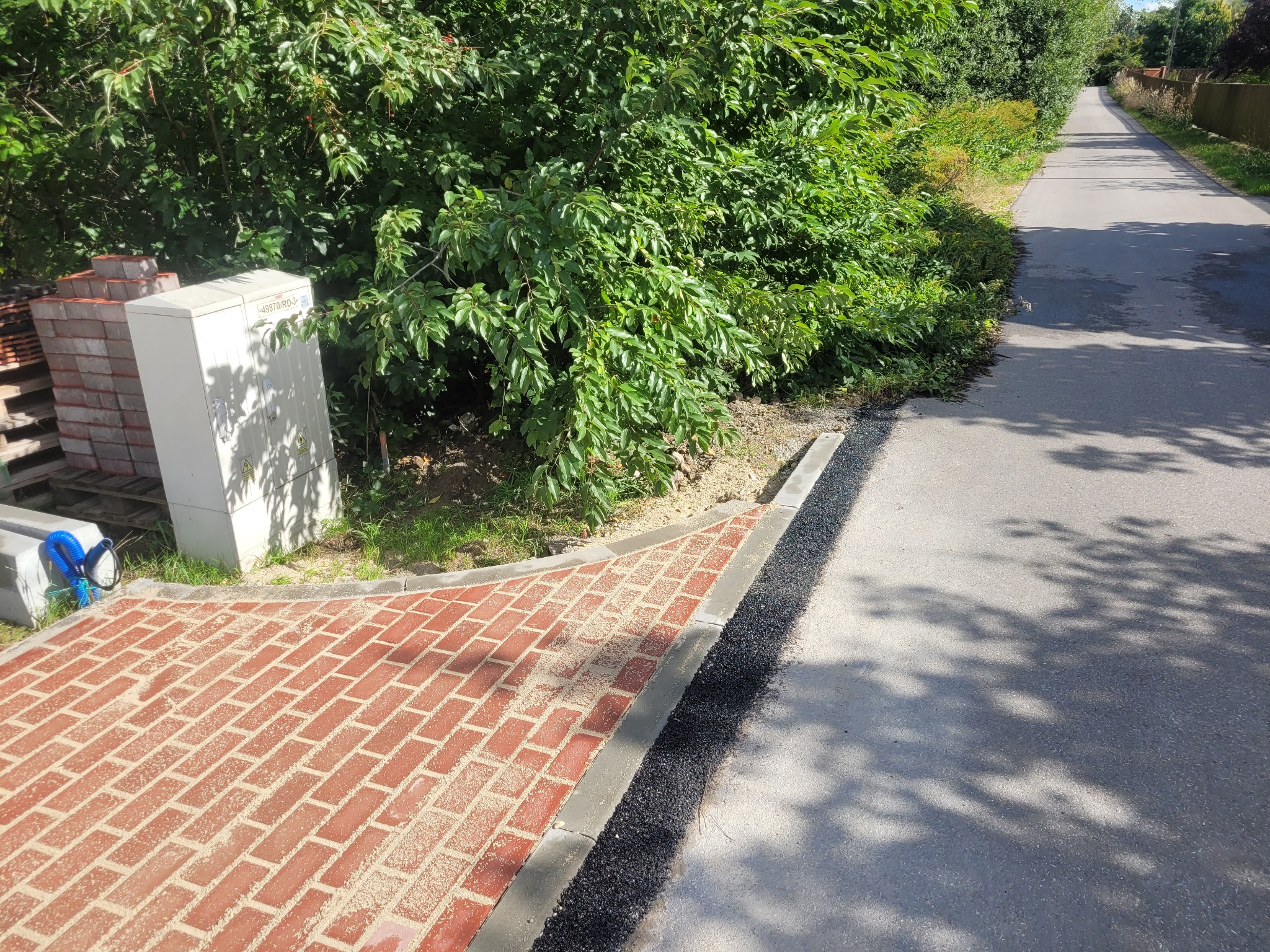 Czerwona kostka brukowa ułożona w jodełkę, obrzeża z szarego betonu, widok na drogę i zieleń. Częściowo zasypana piaskiem. Paleta z kostką w tle.