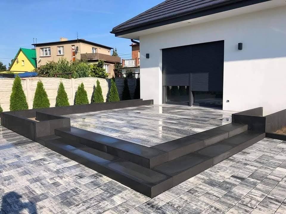 Nowoczesny taras z szaro-białej kostki, z niskimi schodami i podwyższonymi rabatami. Biały budynek w tle, schludne ogrodzenie i tuje.
