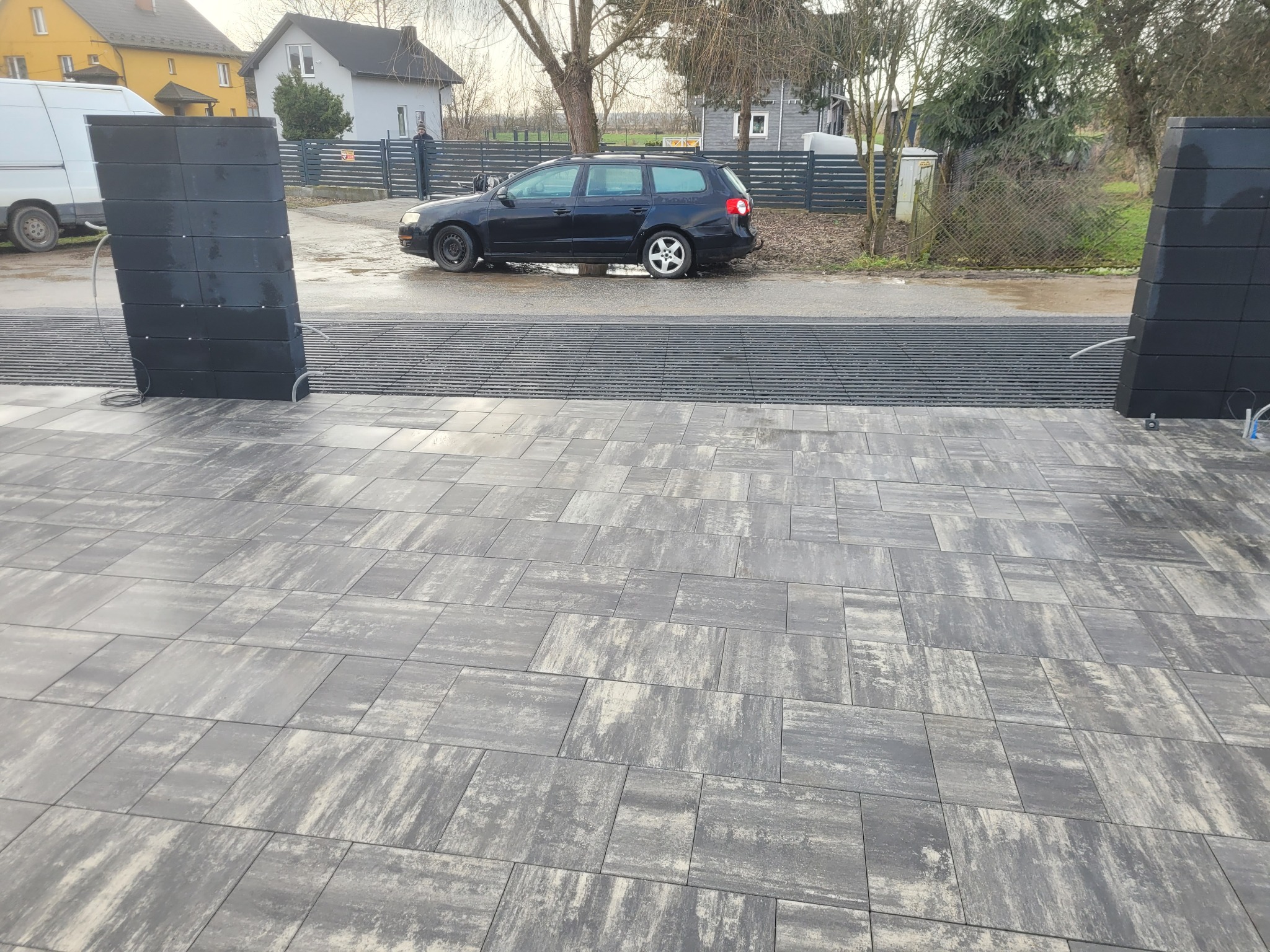 Ułożona kostka brukowa w odcieniach szarości, z widocznym systemem odprowadzania wody. W tle zaparkowany samochód i zabudowa jednorodzinna.