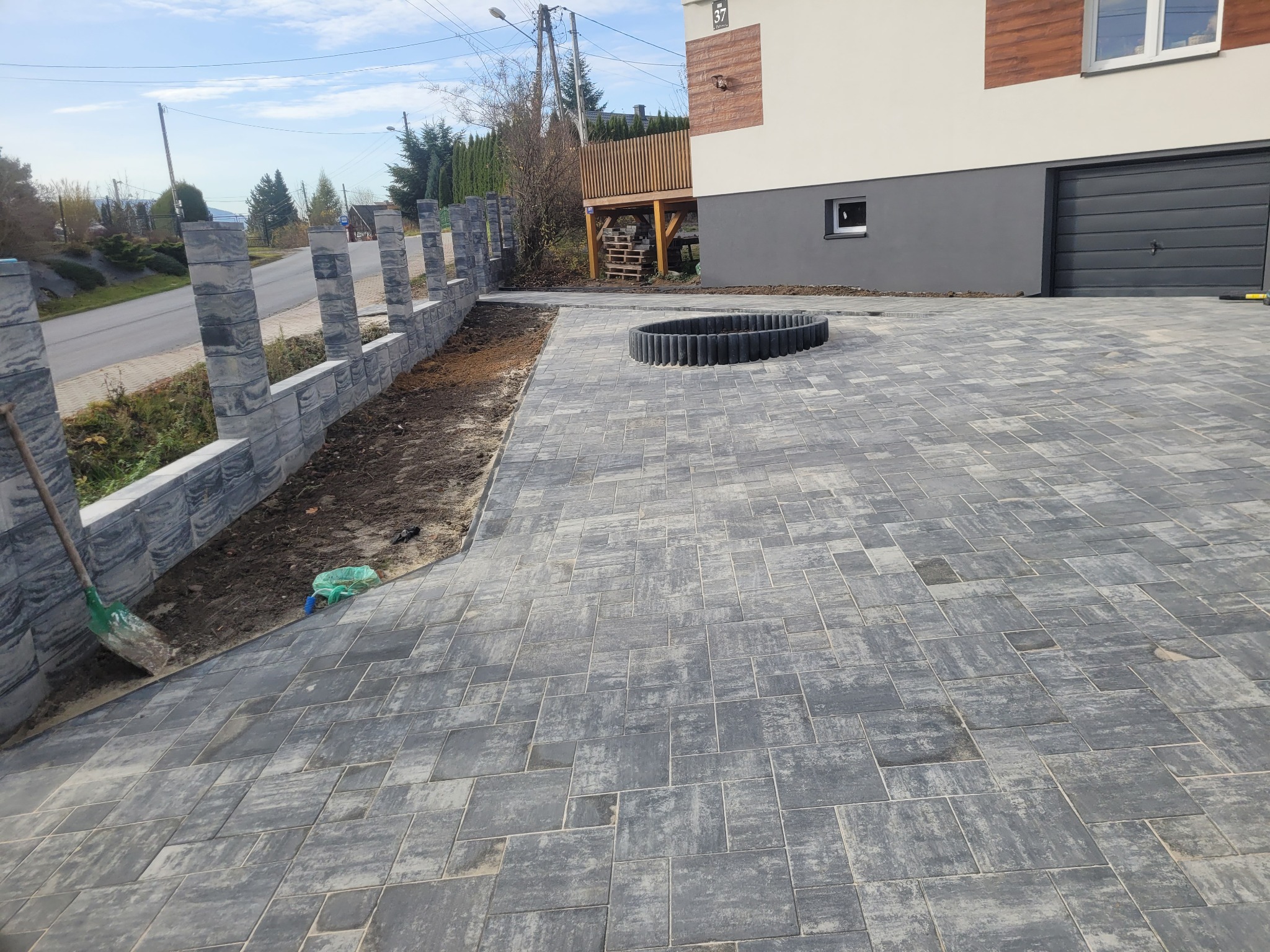 Szara kostka brukowa ułożona na podjeździe, obok ogrodzenie z betonowych elementów. W tle dom z szarą bramą garażową i drewnianym tarasem.