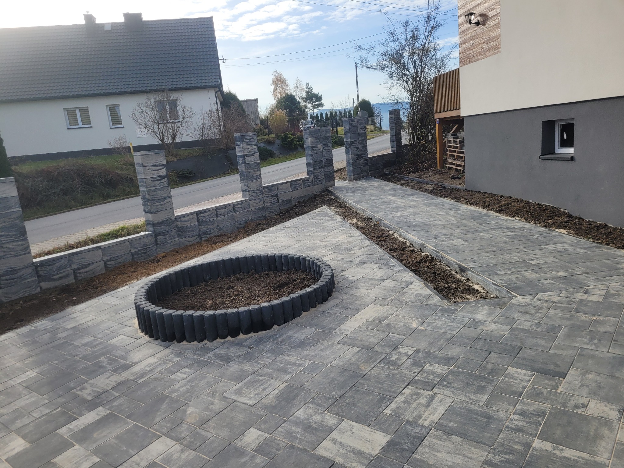 Szara kostka brukowa ułożona na podjeździe z okrągłym klombem i niskim murem z bloków. Widok na dom i ulicę w tle, słoneczny dzień.