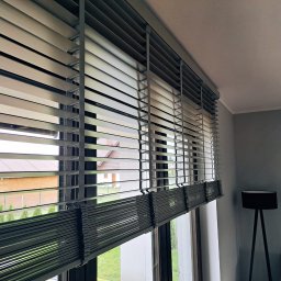 dekoroleta.pl - Zamknięte, szare poziome listwy okienne z widokiem na drewniany dom i zielony trawnik, z lampą na trójnogu w tle.