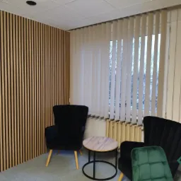 Nowoczesne biuro z pionowymi panelami ściennymi z drewna, białymi żaluzjami w oknie i tapicerowanymi krzesłami w kolorze czarnym i zielonym.