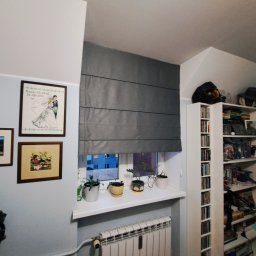 dekoroleta.pl - Szara tkaninowa roleta rzymska zamontowana w oknie, na parapecie doniczki z roślinami, na ścianie obrazy w ramkach, w tle regał z książkami i kolekcją przedmiotów.