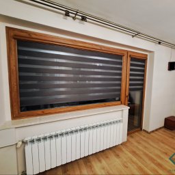 dekoroleta.pl - Okna z drewnianą ramą zabezpieczone roletami dzień-noc, widoczny grzejnik i fragment podłogi z paneli.