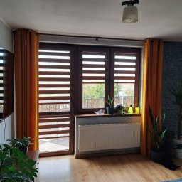 dekoroleta.pl - Jasne wnętrze z oknem przesłoniętym brązowymi żaluzjami poziomymi typu dzień-noc, z pomarańczowymi zasłonami po bokach, roślinami na parapecie i w donicach, oraz dekoracyjną ścianą z szarej cegły...