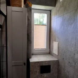 Jasna plisa okienna zamontowana w łazience z szarymi, kamiennymi płytkami, obok szafka łazienkowa i doniczka z rośliną.