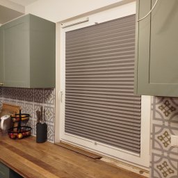 dekoroleta.pl - Szara plisa okienna zamontowana w kuchni, z widocznym blatem, szafkami w odcieniach zieleni i wzorzystymi płytkami nad blatem.