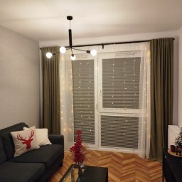 dekoroleta.pl - Salon z szarym narożnikiem, poduszką z motywem renifera i oknem z plisami, ozdobionym lampkami, zasłoniętym częściowo przez oliwkowe zasłony i białą firankę.