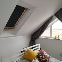 dekoroleta.pl - Pokój na poddaszu z oknem dachowym i plisowaną przesłoną w kolorze czarnym, zasłona w oknie bocznym, dekoracyjne poduszki na łóżku i logo firmy w prawym dolnym rogu.