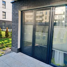 Zewnętrzna moskitiera plisowana w kolorze antracytowym na dwuskrzydłowych drzwiach tarasowych, z widokiem na nowoczesny budynek mieszkalny i starannie utrzymany trawnik.