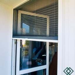 dekoroleta.pl - Zewnętrzne okno z częściowo opuszczonymi plisami w kolorze szarym, widok z balkonu, w tle dekoracyjny element na parapecie.