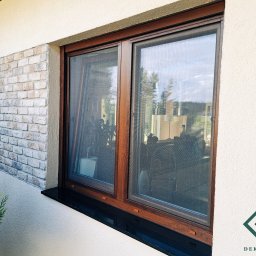 dekoroleta.pl - Brązowe okno z zamontowaną siatką przeciw owadom, widok z zewnątrz, z fragmentem elewacji z cegły i zielonej rośliny.