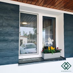 dekoroleta.pl - Okno z zamontowaną siatką przeciw owadom, widok z zewnątrz, na parapecie donica z kwiatami, w tle odbicie samochodu i budynku.