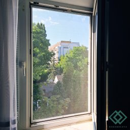 dekoroleta.pl - Widok z okna zabezpieczonego siatką przeciw owadom, na zewnątrz zieleń drzew i budynek wielorodzinny, po lewej stronie okna firanka, po prawej ciemne wnętrze z widocznym logo firmy.