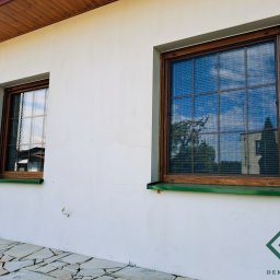 dekoroleta.pl - Dwa okna z drewnianą ramą i poziomymi przesłonami w białym budynku, widoczne odbicie drzewa w szybie, na ścianie logo firmy.