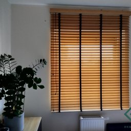dekoroleta.pl - Jasne, drewniane żaluzje poziome z czarnymi taśmami na oknie, częściowo zasłonięte, z widocznym grzejnikiem pod oknem i rośliną doniczkową z lewej strony