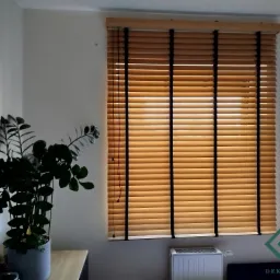 Jasne, drewniane żaluzje poziome z czarnymi taśmami na oknie, częściowo zasłonięte, z widocznym grzejnikiem pod oknem i rośliną doniczkową z lewej strony