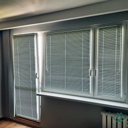 dekoroleta.pl - Zamknięte, poziome, aluminiowe osłony okienne w szarym pomieszczeniu, zamontowane w oknach z białymi ramami.