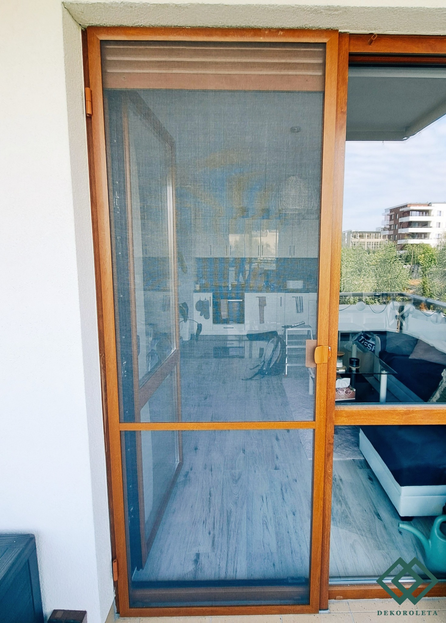 Drewniana rama z siatką na owady, przesuwana wzdłuż balkonu, z widokiem na wnętrze mieszkania i zabudowę miejską w tle.