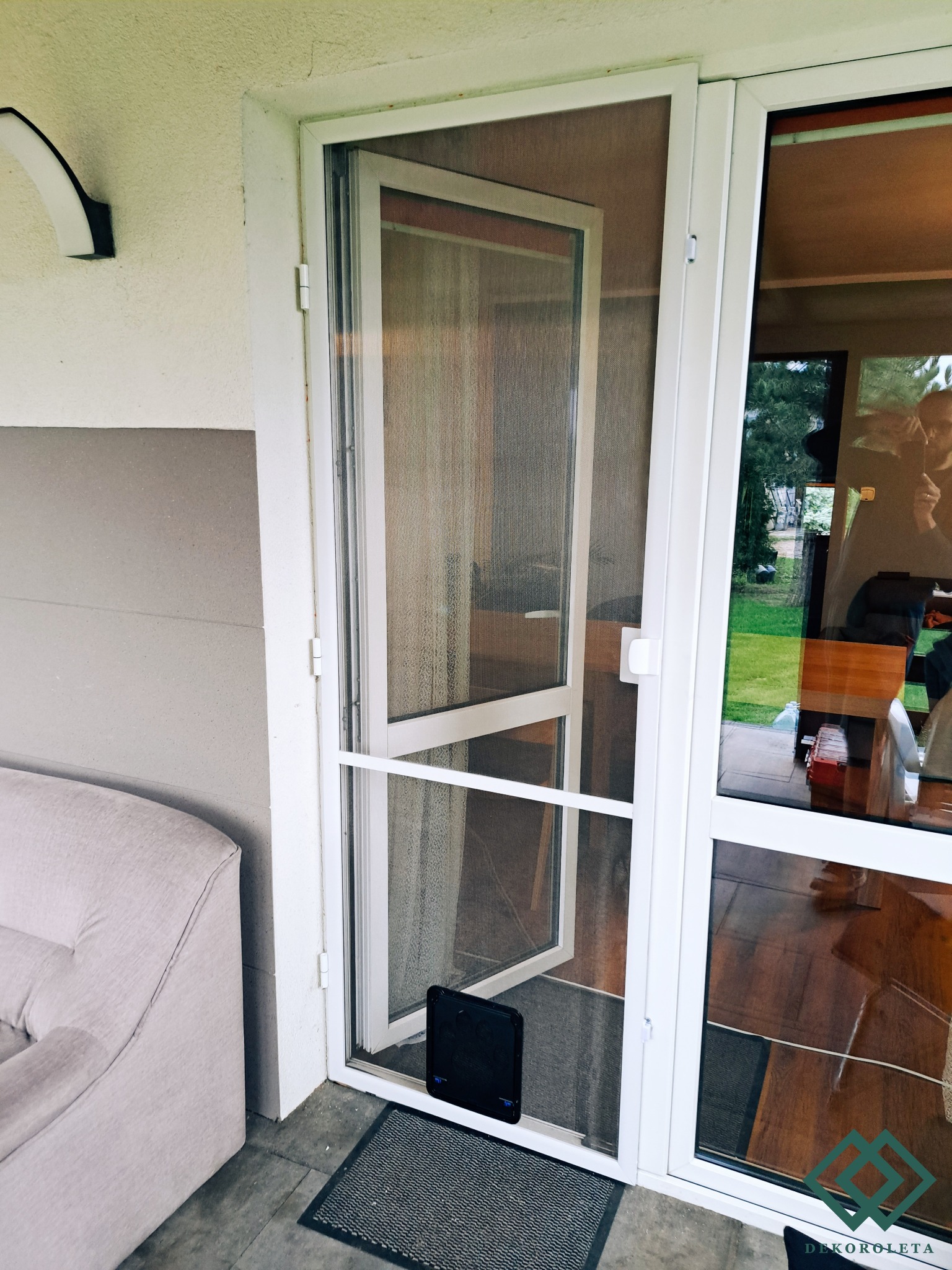 Otwarta, biała rama drzwiowa z siatką przeciw owadom, zamontowana przy drzwiach tarasowych z widocznym przejściem dla zwierząt w dolnej części, na zewnątrz widoczny trawnik.