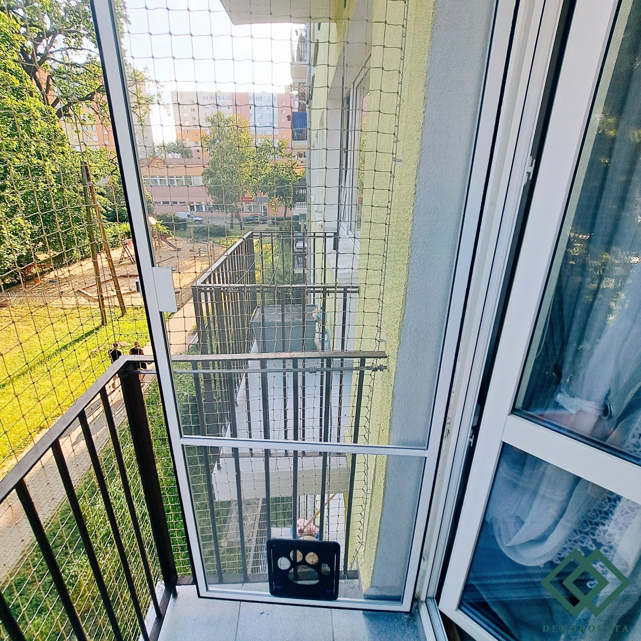 Zabezpieczenie balkonu siatką ochronną dla kota, widok z wewnątrz mieszkania na balkon i plac zabaw, czarna kuweta z wyciętym wzorem łapy kota na podłodze balkonu.