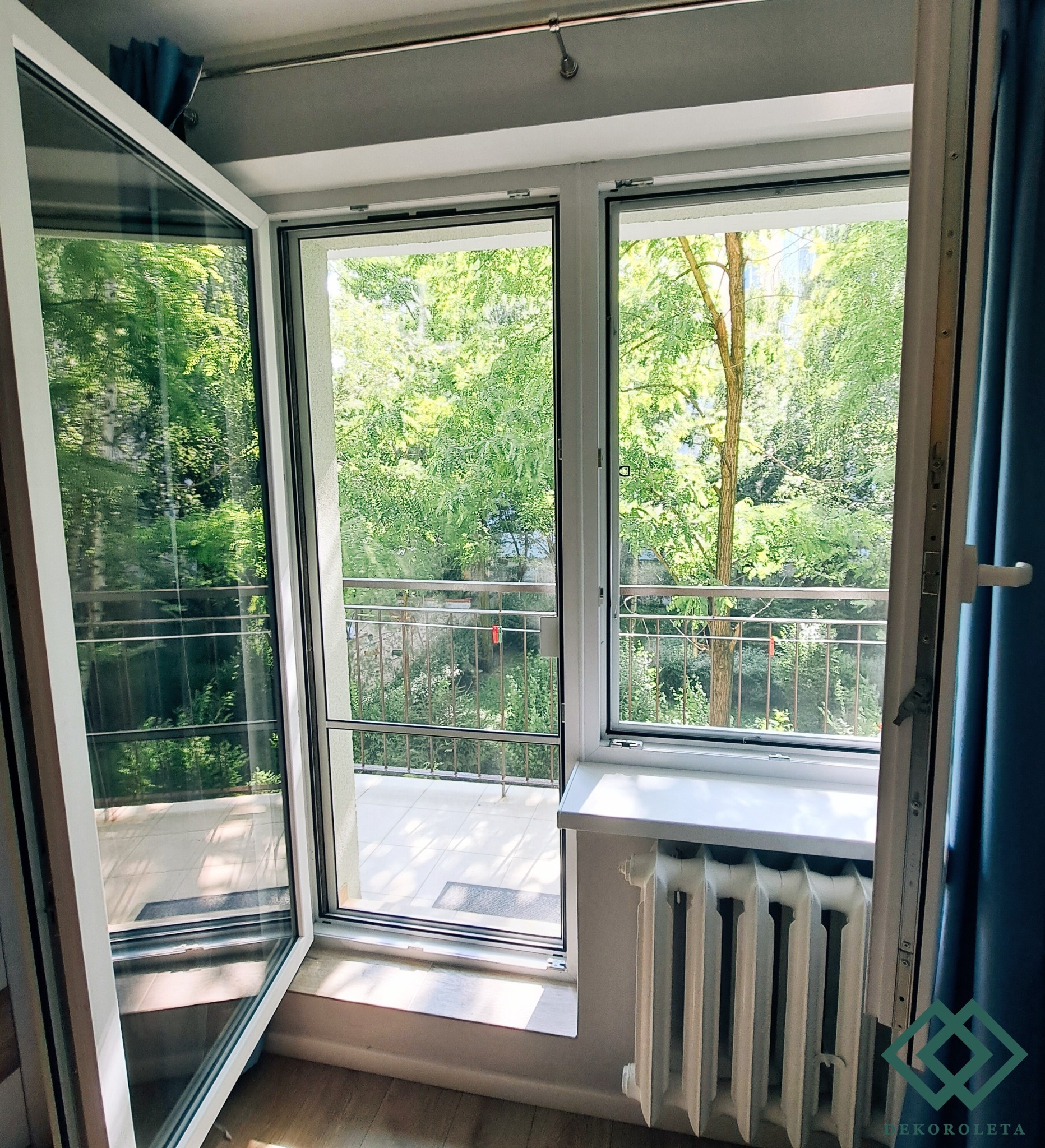 Otwarty balkon z widokiem na zieleń, zabezpieczony moskitierą w białej ramie okiennej, widoczny fragment grzejnika i zasłony.