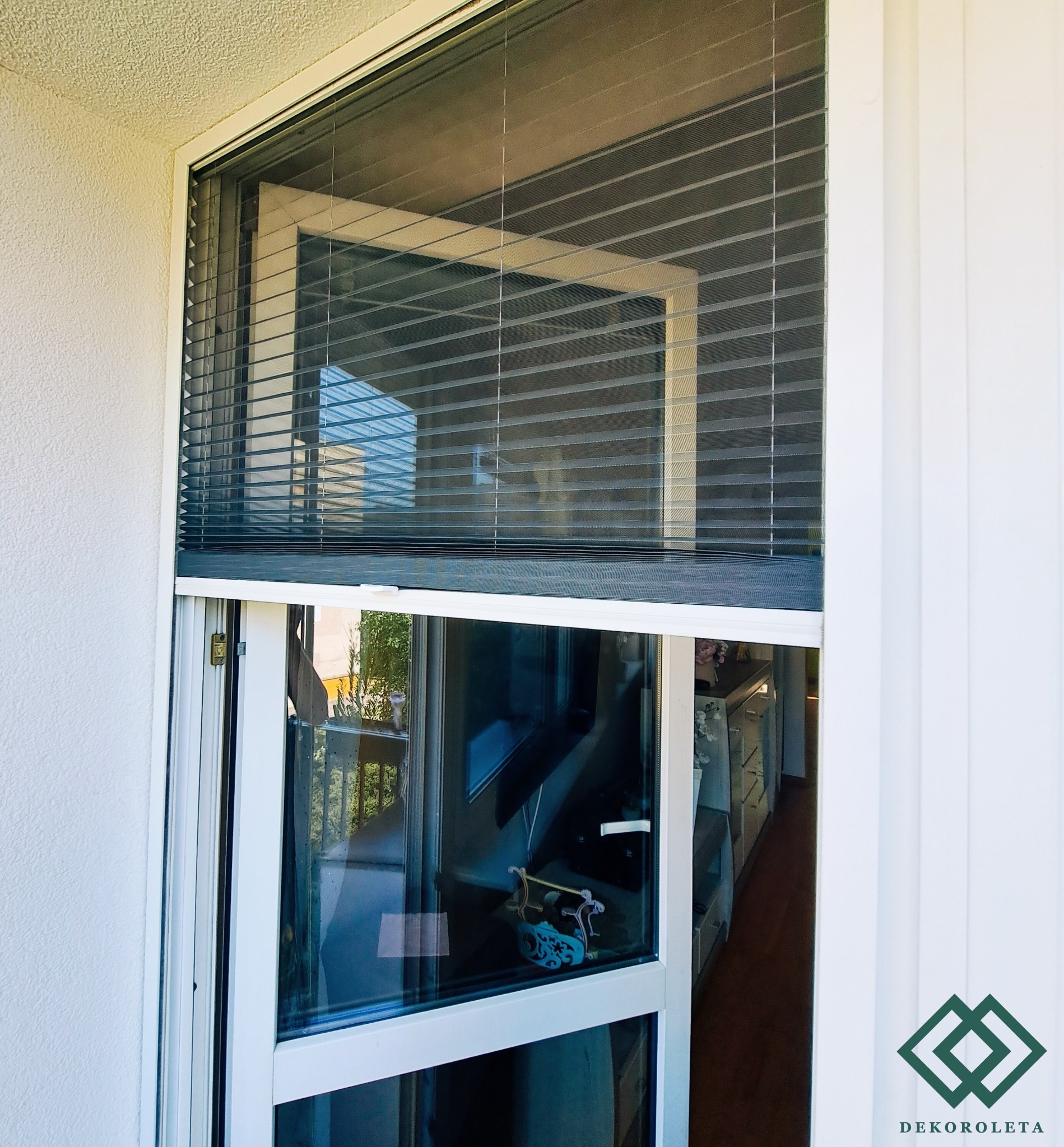 Zewnętrzne okno z częściowo opuszczonymi plisami w kolorze szarym, widok z balkonu, w tle dekoracyjny element na parapecie.