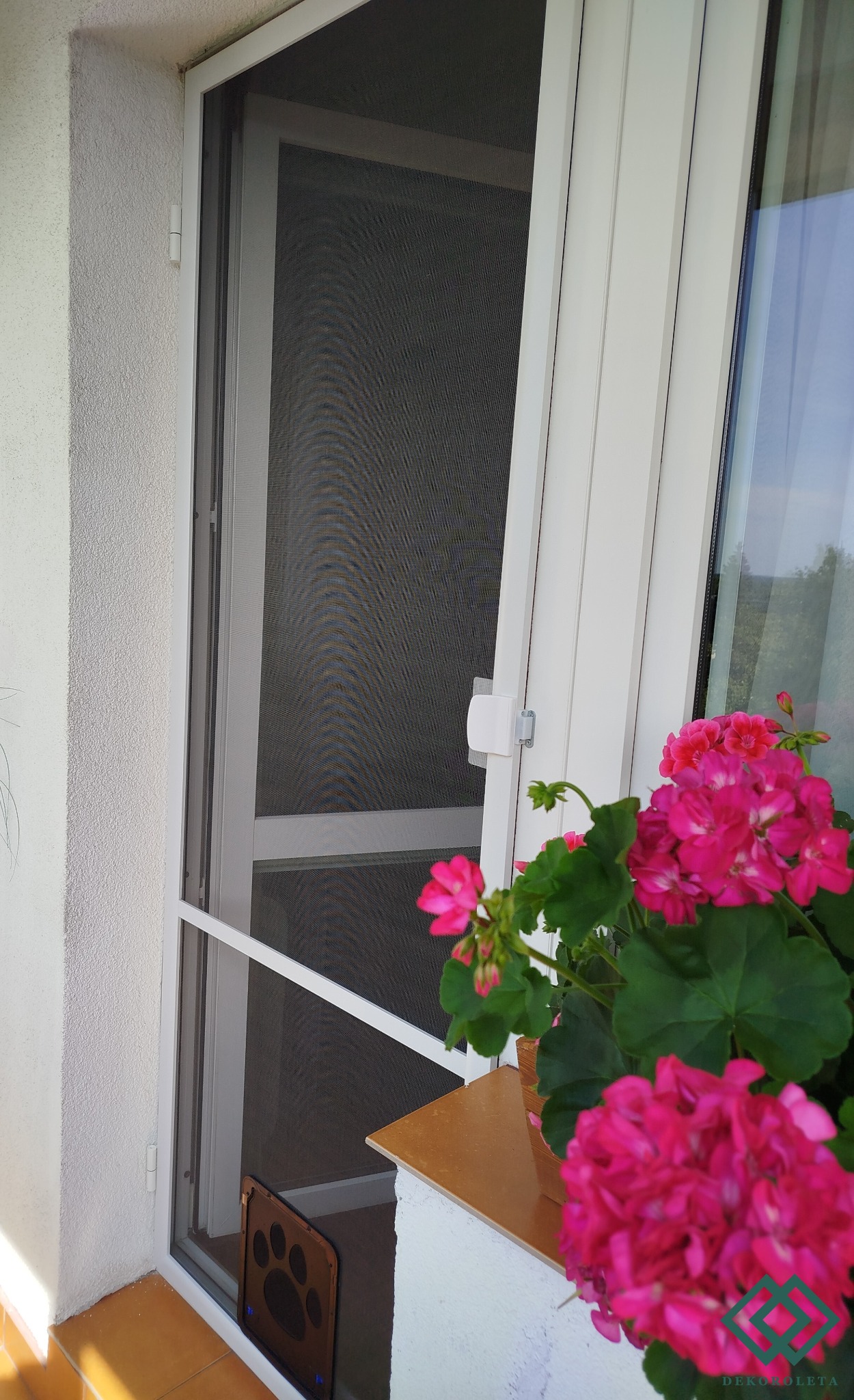 Biała rama drzwi balkonowych z siatką przeciw owadom, widoczna z boku, z doniczką z różowymi pelargoniami na parapecie i naklejką z motywem psiej łapy na dole ramy.