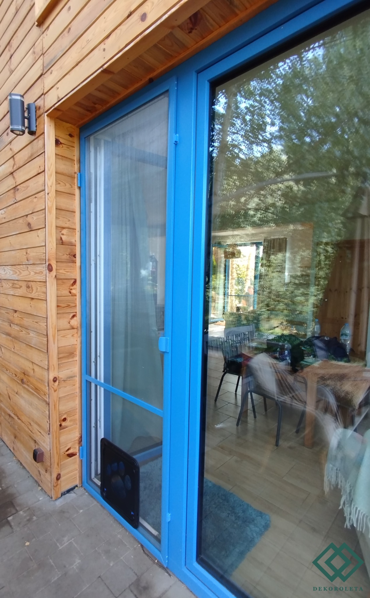 Błękitna moskitiera z klapą dla zwierząt, zamontowana w drzwiach tarasowych w Toruniu, widoczna na tle drewnianej elewacji domu.