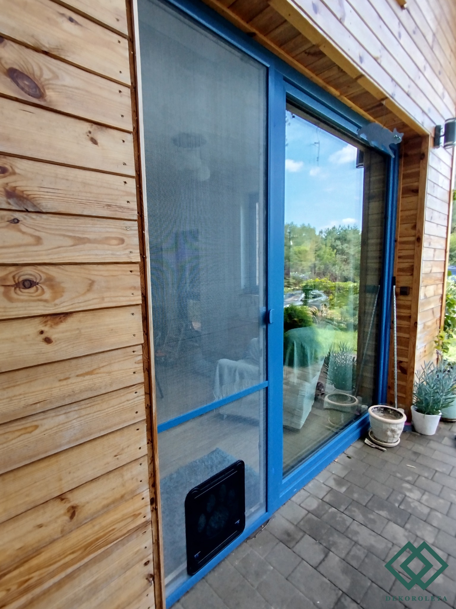 Zewnętrzny widok na okno przesuwne z niebieską ramą i siatką przeciw owadom oraz klapką dla zwierząt w dolnej części, na tle drewnianej elewacji domu.