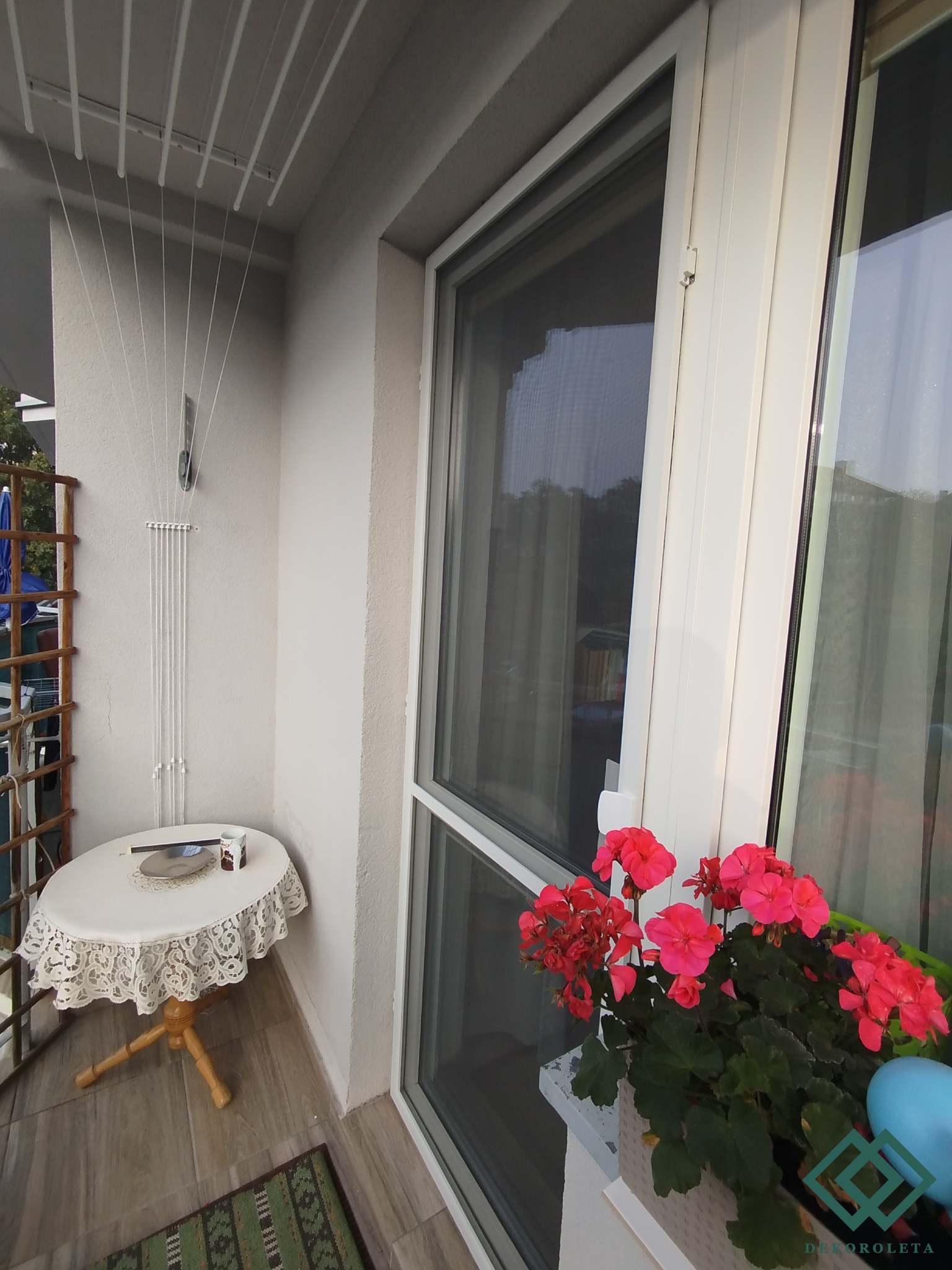 Zabezpieczony siatką balkon z suszarką sufitową, stolikiem z koronkowym obrusem i pelargoniami w doniczce na parapecie.