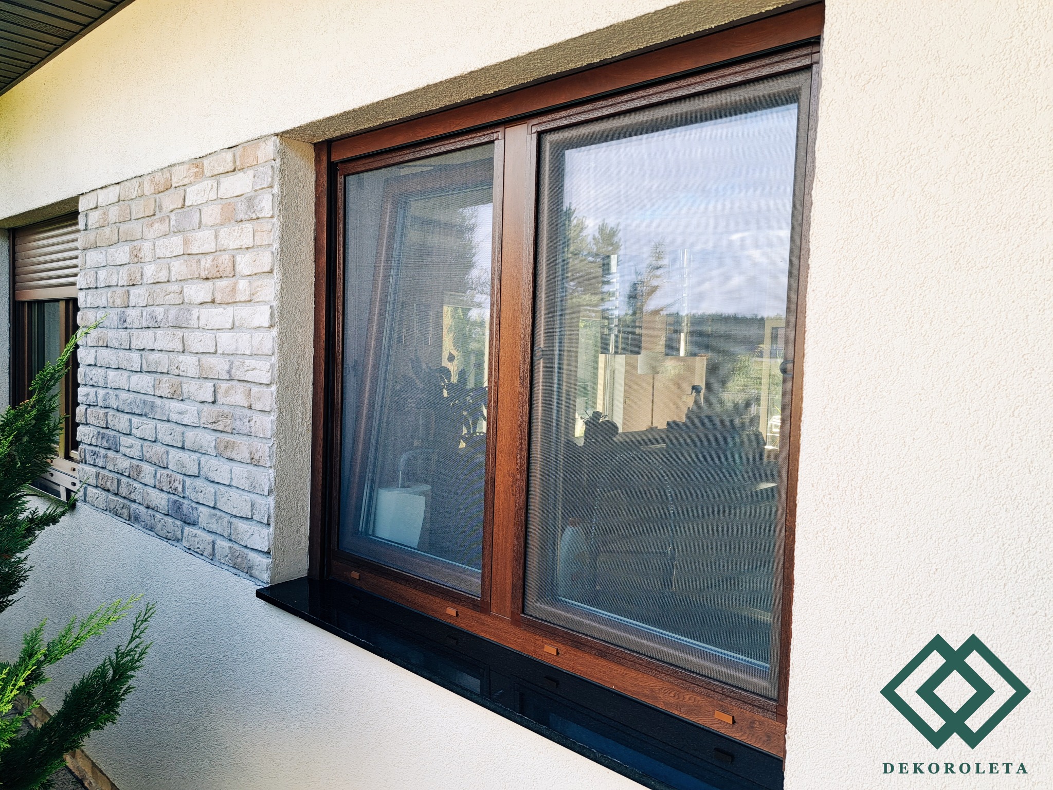 Brązowe okno z zamontowaną siatką przeciw owadom, widok z zewnątrz, z fragmentem elewacji z cegły i zielonej rośliny.