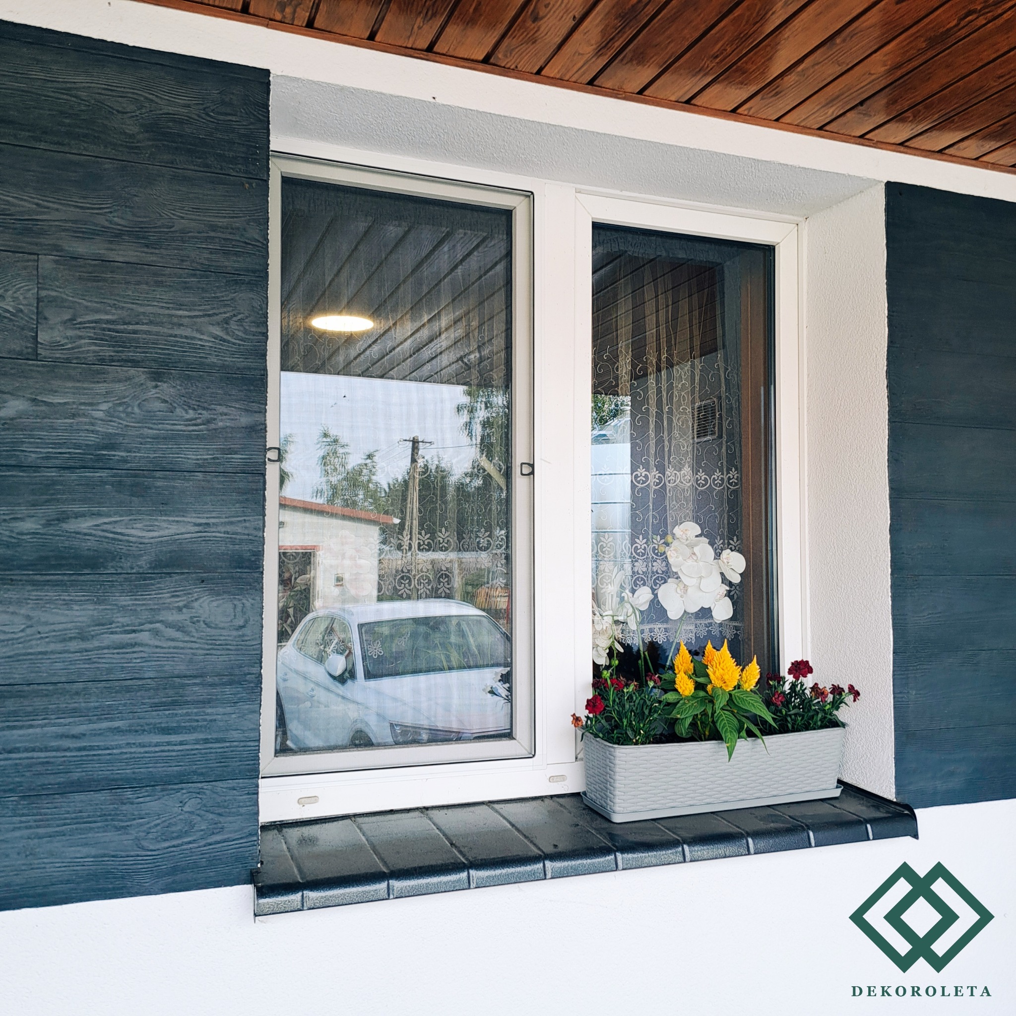 Okno z zamontowaną siatką przeciw owadom, widok z zewnątrz, na parapecie donica z kwiatami, w tle odbicie samochodu i budynku.