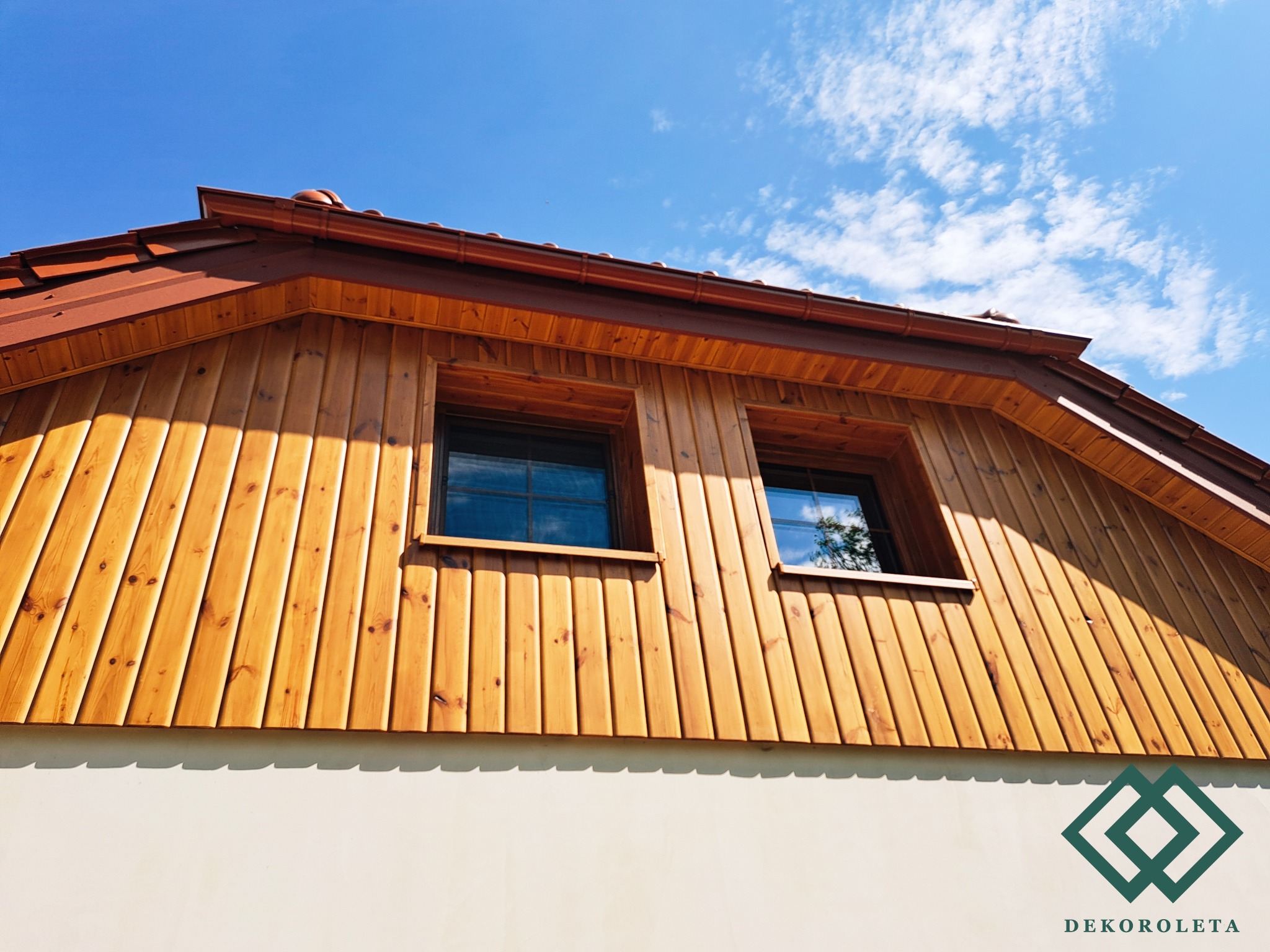 Drewniana fasada poddasza z dwoma oknami, brązowy dach i błękitne niebo z chmurami w tle.