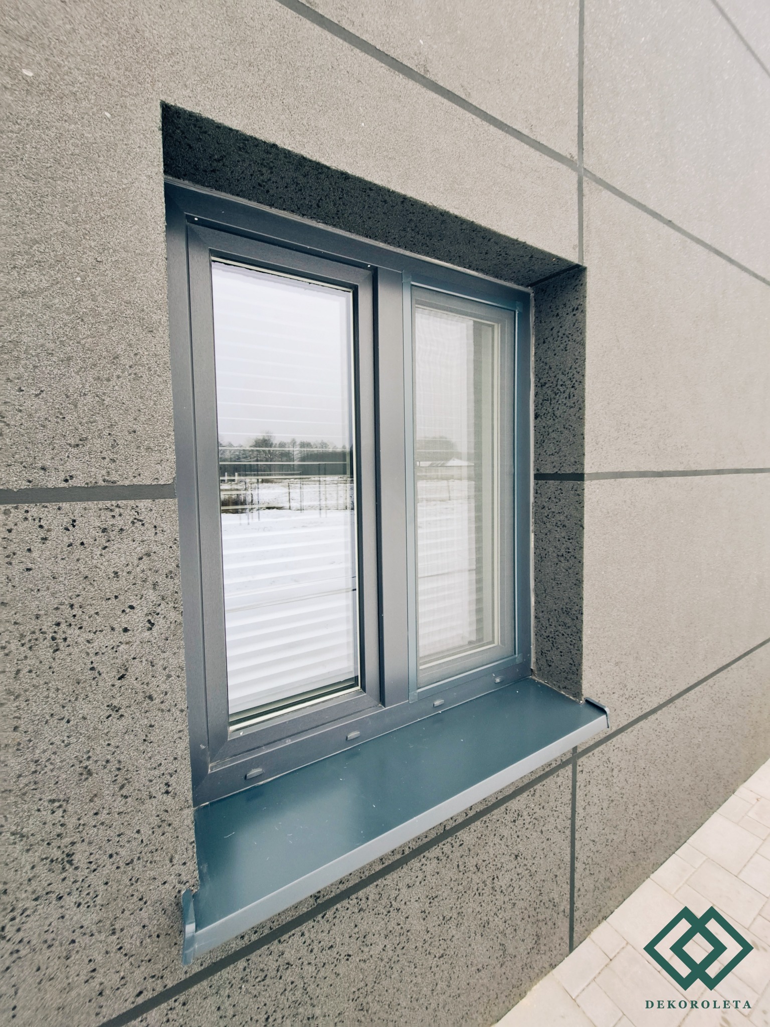 Szare okno z poziomymi przesłonami wewnętrznymi w elewacji z szarego betonu architektonicznego.