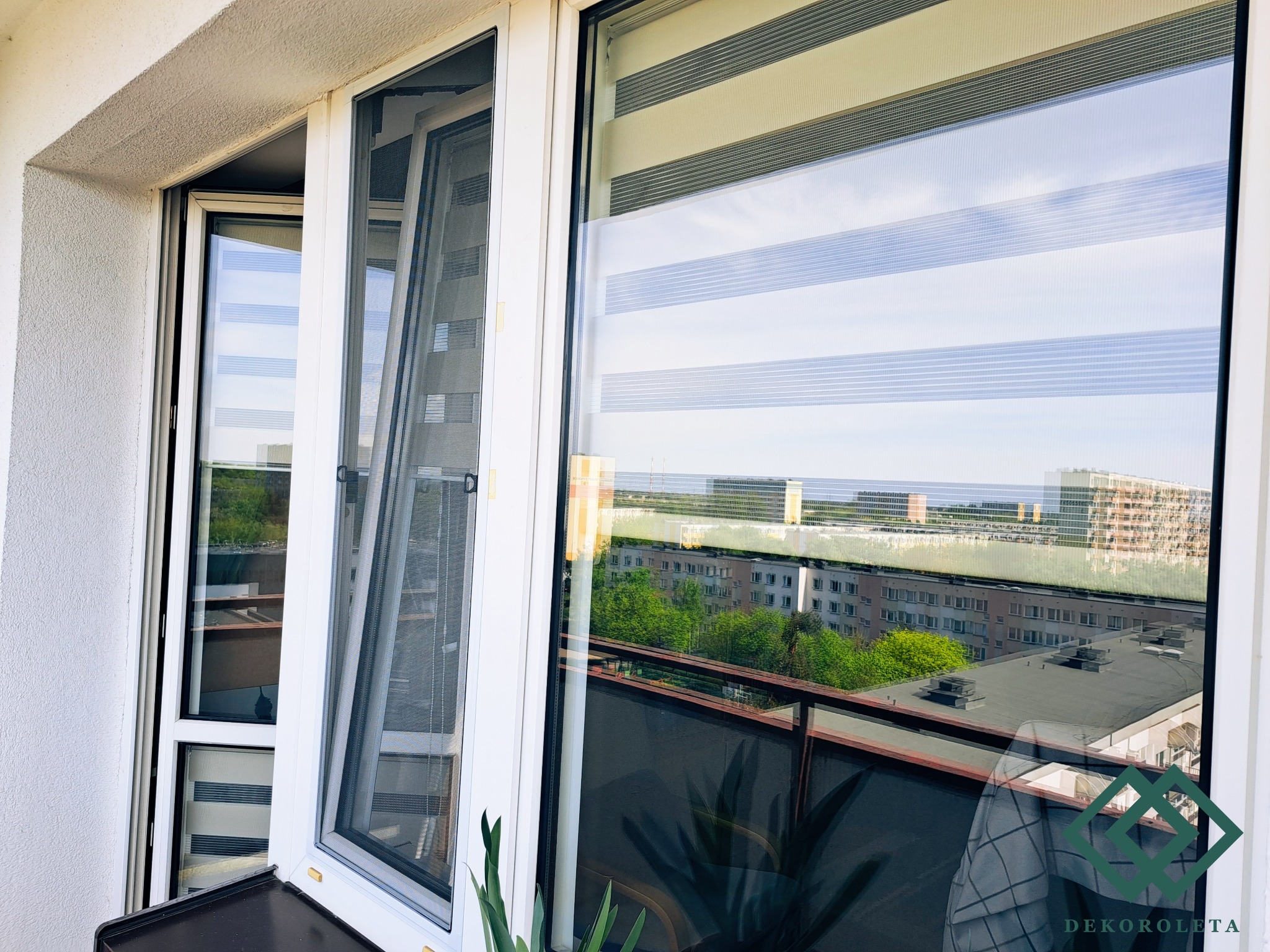 Białe okno balkonowe z zamontowaną moskitierą i roletą dzień-noc, widok na osiedle z balkonu.