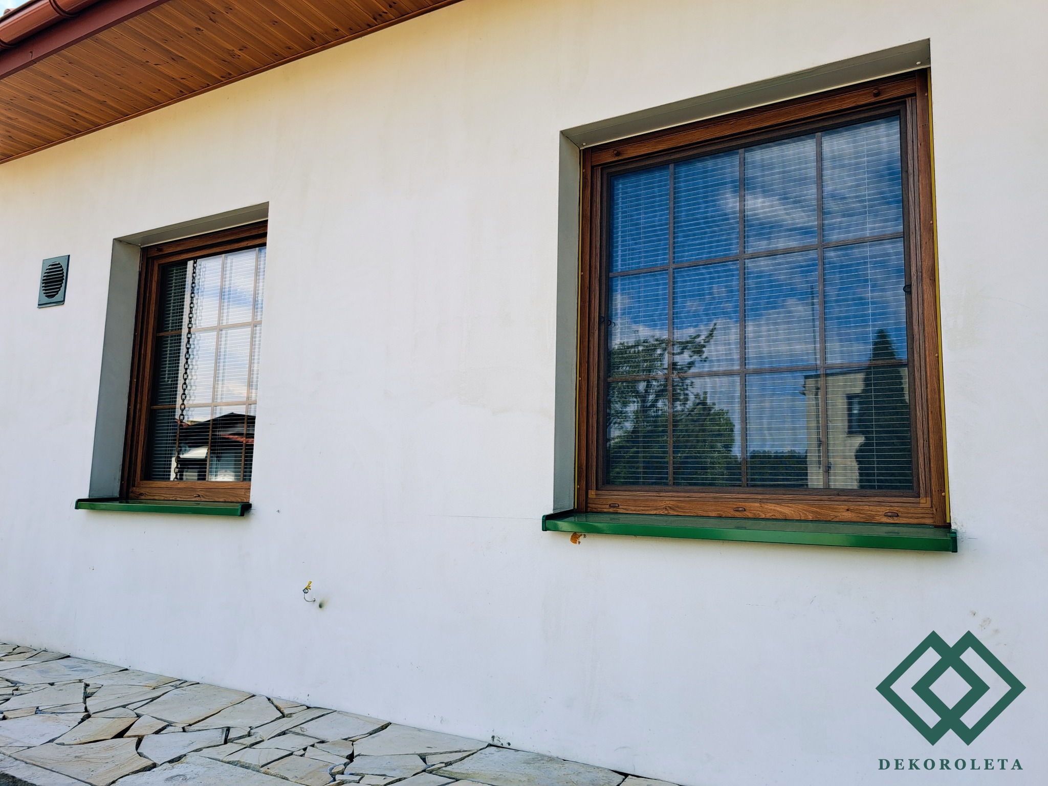 Dwa okna z drewnianą ramą i poziomymi przesłonami w białym budynku, widoczne odbicie drzewa w szybie, na ścianie logo firmy.