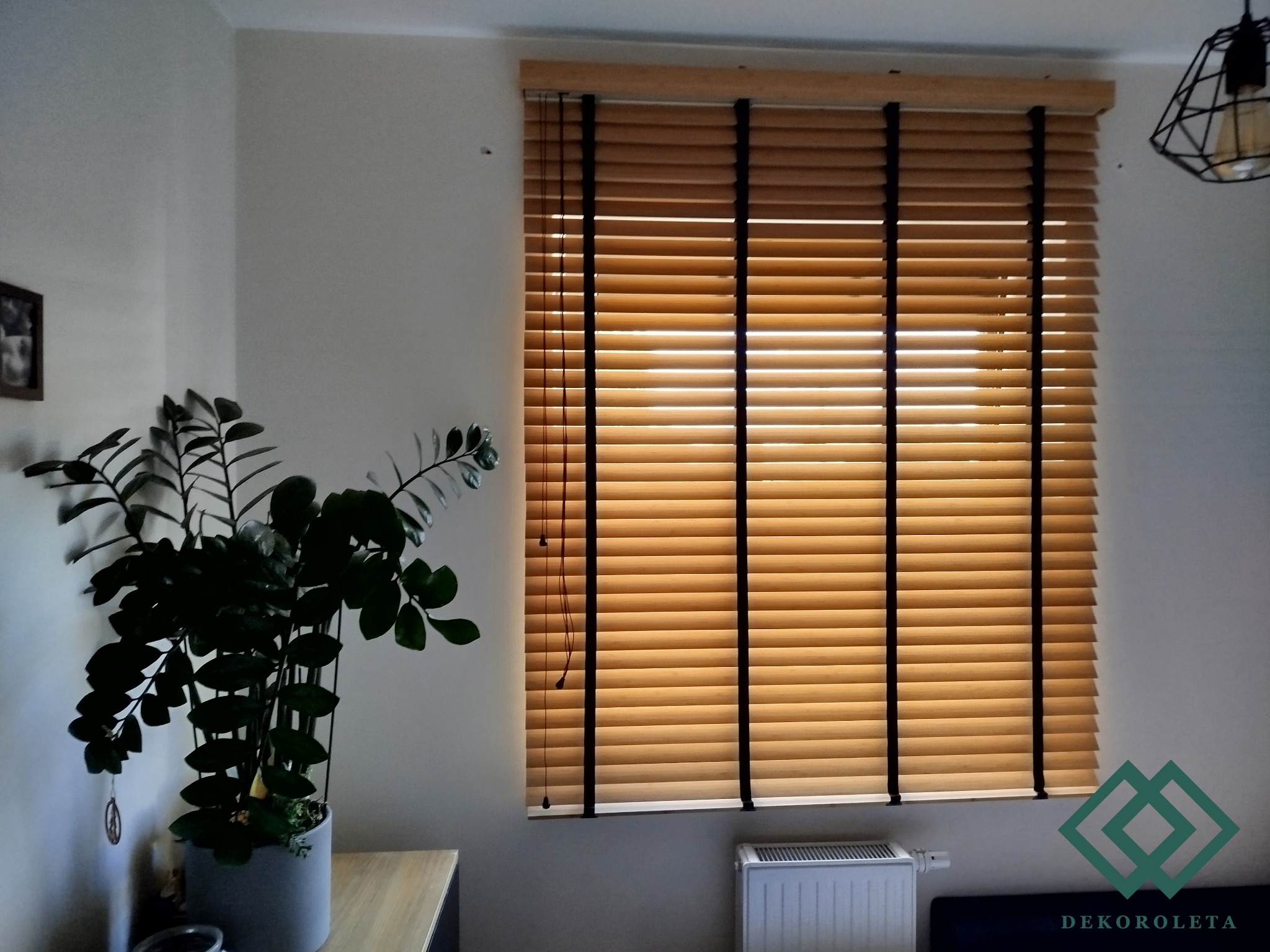 Jasne, drewniane żaluzje poziome z czarnymi taśmami na oknie, częściowo zasłonięte, z widocznym grzejnikiem pod oknem i rośliną doniczkową z lewej strony