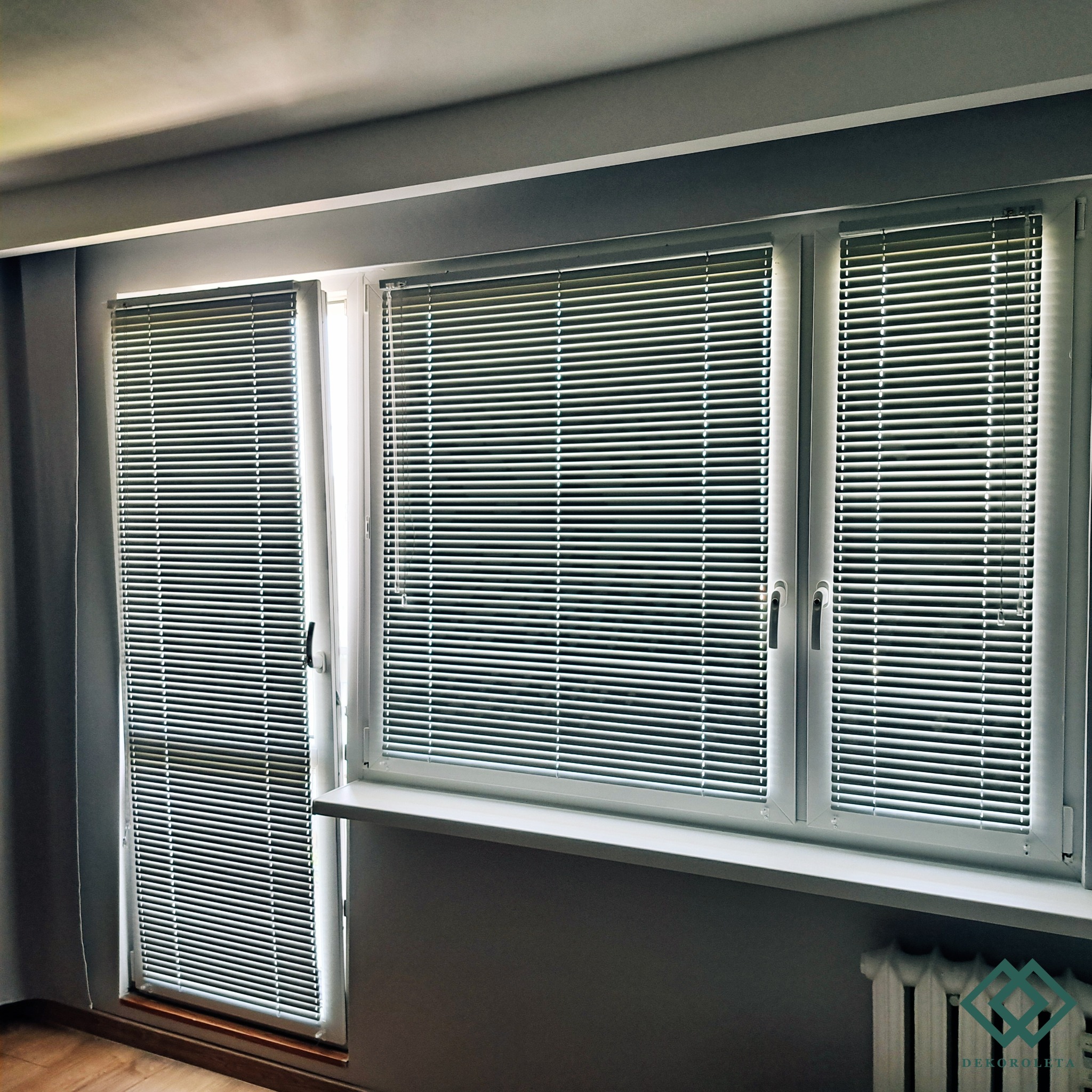 Zamknięte, poziome, aluminiowe osłony okienne w szarym pomieszczeniu, zamontowane w oknach z białymi ramami.